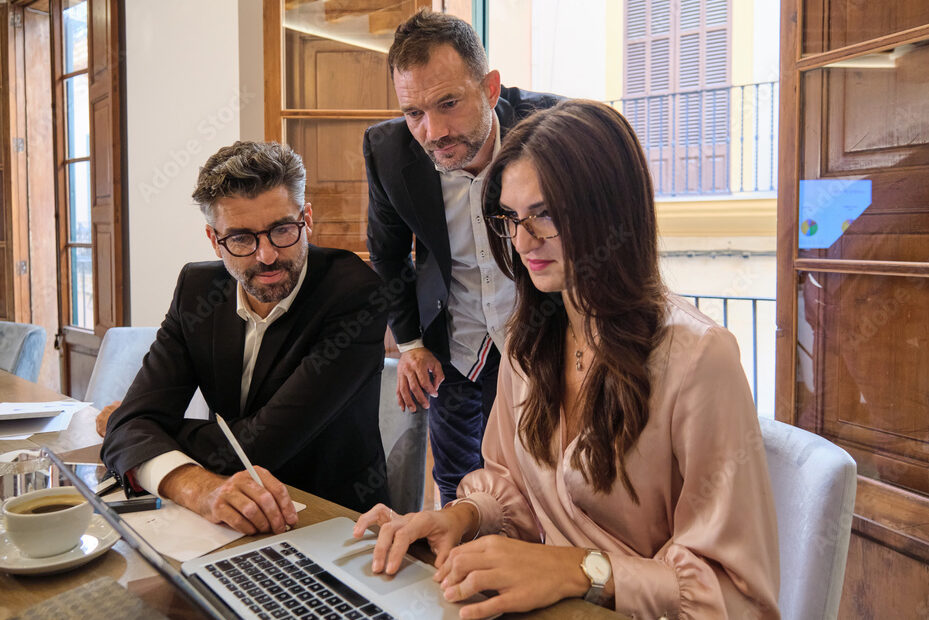 Group of modern adult coworkers checking information on laptop while sitting at table and discussing business plans during meeting in workspace