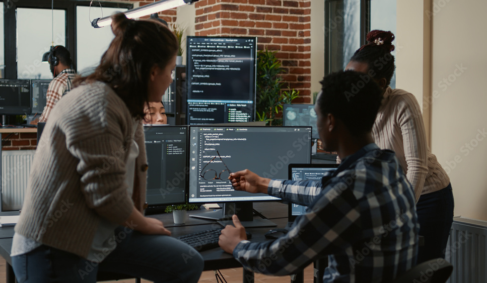 Programer sitting on desk discussing with mixed team of software developers about artificial intelligence innovation. Programmers doing teamwork looking at running machine learning algorithms.