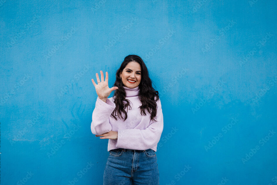 Smiling young beautiful woman showing number 5 in front of blue wall